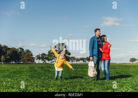 Verspielter kleiner Kind läuft auf der grünen Wiese, spielt in der Nähe von Eltern, die sich umarmen, Blick in die Ferne mit durchdachten Ausdrücke. Familie hat zu Fuß Stockfoto