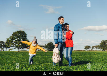 Junges Paar in der Liebe stehen auf grünem Gras, umarmen und nachdenklich. Verspielte kleine Kind Freiheit genießt, läuft auf Feld, spielt in der Nähe von ihren Eltern, hat Stockfoto