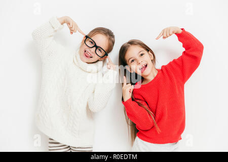 Fröhliches Mädchen, die in guter Stimmung, Verschleiß übergroße Pullover und Brillen, machen coole Zeichen, gemeinsam gegen den weißen Hintergrund darstellen. Gerne weibliche Kinder Stockfoto