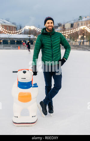 Außenaufnahme der froh, bärtigen Mann trägt grüne Jacke und Hut, steht in der Nähe von Skate Beihilfen wie der Versuch, Skating Skills zu bekommen, steht auf Eisbahn, hat entert Stockfoto