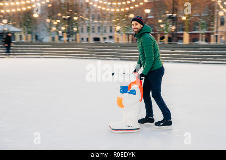 Gerne männliche Skater, auf der Eisbahn, verwendet skate Beihilfe als versucht, im Gleichgewicht zu sein, schaut gerne beiseite, stellt auf dem Eis. Fröhlicher Mann in warm gekleidet c Stockfoto