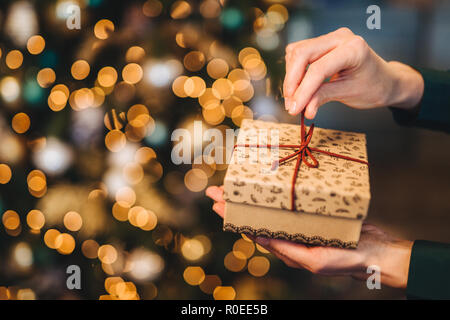 Nicht erkennbare Frau wrappes Geschenkbox über Weihnachten Baum mit leuchtenden Lichter und Kränze. Dekoriert präsentieren, in Frau ein Hände. Urlaub und celeb Stockfoto