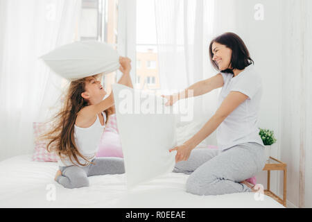 Innen- schuss von angenehmen suchen europäische Weibchen hat Freude zusammen mit ihrer kleinen Tochter, haben, Kissen, Kampf im Schlafzimmer, Pose in der gemütlichen geräumigen hellen Ro Stockfoto