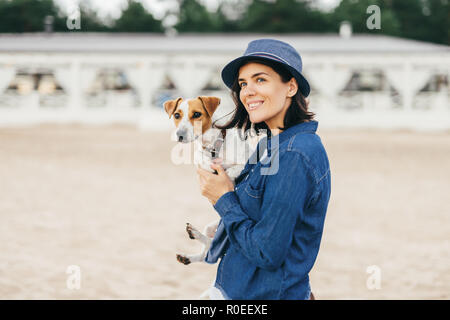 Schöne Frau trägt modischen Hut und Jeanshemd, Spaziergang mit ihrem Hund im Freien, unbekannte Orte finden, schaut nachdenklich in die Ferne, hat h Stockfoto