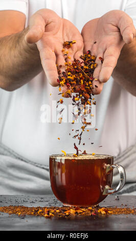 Der Mann am Tisch gießt aus der Höhe eine Mischung aus Obst und Kräuter in ein Glas roten Tee Stockfoto