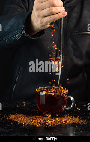 Der Mann am Tisch gießt aus der Höhe eine Mischung aus Obst und Kräuter in ein Glas roten Tee Stockfoto