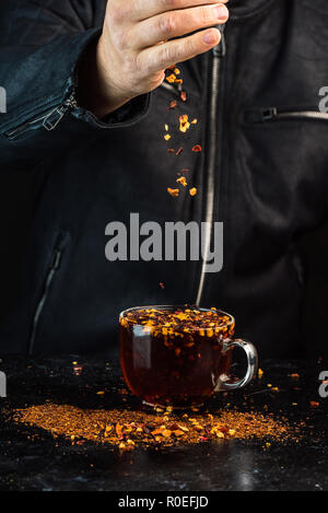 Der Mann am Tisch gießt aus der Höhe eine Mischung aus Obst und Kräuter in ein Glas roten Tee Stockfoto