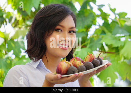 Junge schöne asiatische Frau Lächeln, während Sie einen Teller mit frischen Feigen Früchte. Ein gutes Konzept Bild für Obst essen Förderung gesunder Nahrung und Essen