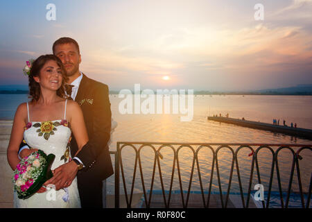 Ein junges Paar, die am Tag ihrer Hochzeit posiert bei Sonnenuntergang liegen auf einem Balkon über dem Hafen von Syrakus in Sizilien. Stockfoto