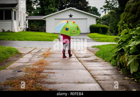 Ein 4-jähriges Mädchen Spaziergänge im Regen mit einem Schirm und tragen regen Stiefel. Stockfoto