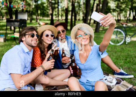 Lächelnd multikulturellen Freunde mit Bier unter selfie auf dem Smartphone während im Sommer Park ruhenden Stockfoto