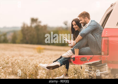 Seitenansicht des glückliches junges Paar mit Gadgets während der Sitzung in Kofferraum während der Reise Stockfoto