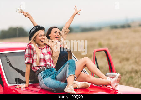 Glückliche junge Freundinnen sitzen auf Auto Motorhaube mit erhobenen Händen und Frieden schildern Stockfoto