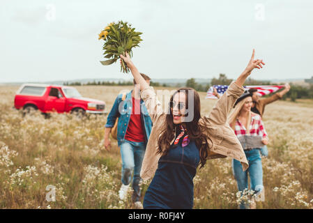 Glückliche junge Frau mit Blumenstrauß und wandern durch Feld mit Freunden während der Reise Stockfoto