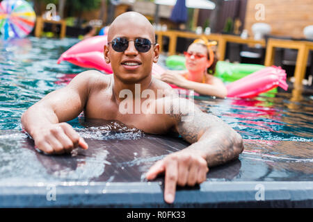 Jungen tätowierten Mann in Sonnenbrille an der Kamera, während seine Freundin ruht auf aufblasbare Matratze im Pool Stockfoto
