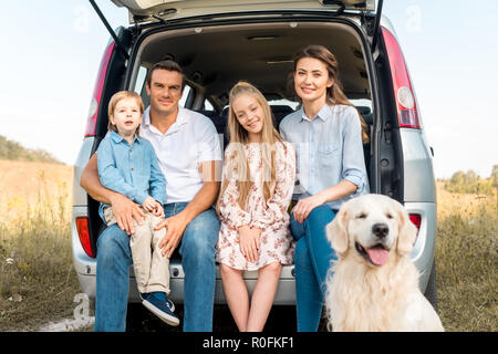 Schöne junge Familie mit Retriever Hund im Kofferraum sitzen und Kamera im Feld Stockfoto