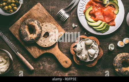 In der hälfte Bagel Brötchen auf hölzernen ausnehmen Board mit Brotmesser und Sandwich Zutaten: Lachs, Avocado, Hummus und Wachteleier, Ansicht von oben Stockfoto