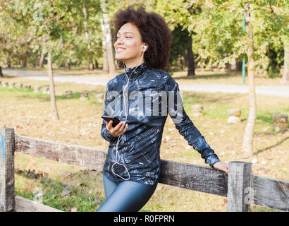Attraktive sportliche Frau entspannen Sie sich nach dem Training Musik im Park, stützte sich auf hölzernen Zaun in der Natur und an der Seite Stockfoto