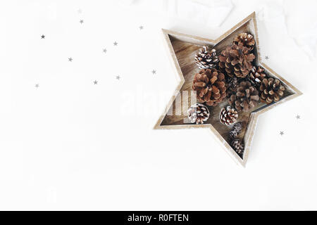 Weihnachten gestaltete Tabelle Zusammensetzung. Kiefer und lärche Kegel in sternförmigen Holz- fach, silber Konfetti und Seidenband auf weißem Hintergrund. Flach, Ansicht von oben. Winter patttern. Stockfoto
