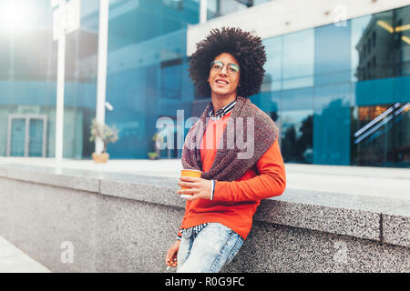 Junge curly Mixed-race Junge trinkt Kaffee auf der Straße vor dem Hintergrund von Bürogebäuden. Jugend Kultur und Mode. Student auf eine Pause zwisch Stockfoto