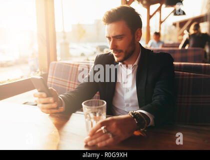 Tausendjährige Geschäftsmann in einem Cafe sitzen an einem Tisch und auf den Bildschirm seines Handys. Junge erfolgreiche stilvollen Mann ruht in einem Sommer Cafe Stockfoto