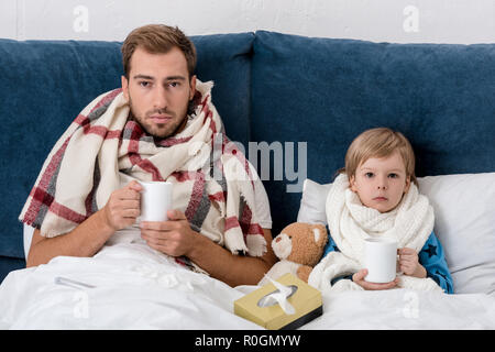 Kranken Vater und Sohn in Schals mit Tassen heißes Getränk im Bett sitzen und Kamera Stockfoto