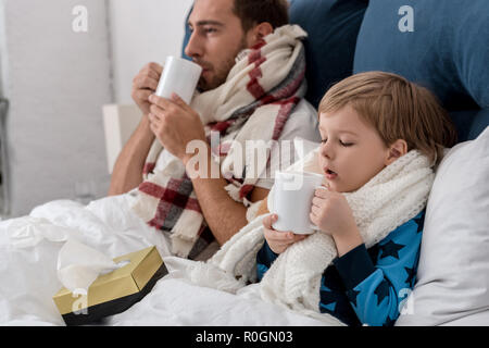 Kranken Vater und Sohn in Schals mit Tassen heißes Getränk im Bett sitzen Stockfoto