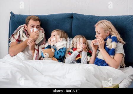 Kranke junge Familie blasen Nasen mit Servietten zusammen, während im Bett lag, und bei jeder anderen Suchen Stockfoto