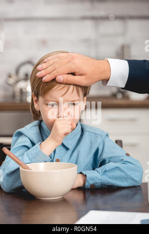 Schoß von Vater Kontrolle der Temperatur von kranken Sohn mit Hand beim Frühstück 7/8 Stockfoto