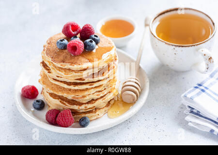 Stapel von gesunden oat Pfannkuchen mit Beeren, Honig und Tasse grünen Tee auf Weiß konkreten Hintergrund, selektive konzentrieren. Leckeres und gesundes Frühstück Stockfoto