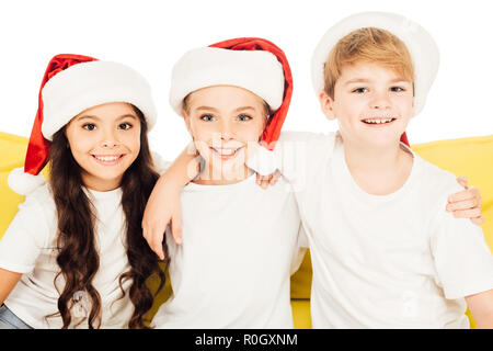 Portrait von lächelnden adorable Kinder in santa Hüte sitzen auf gelbes Sofa und Blick auf Kamera isoliert auf weißem Stockfoto