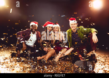 Fröhlich multikulturellen Freunde in santa Hüte holding Champagner Gläser und sitzen auf Konfetti Stockfoto