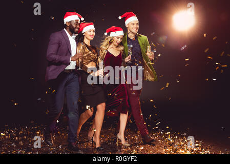 Glamouröse multiethnischen Freunde in santa Hüte holding Champagner Gläser und gehen auf Konfetti mit Hintergrundbeleuchtung Stockfoto