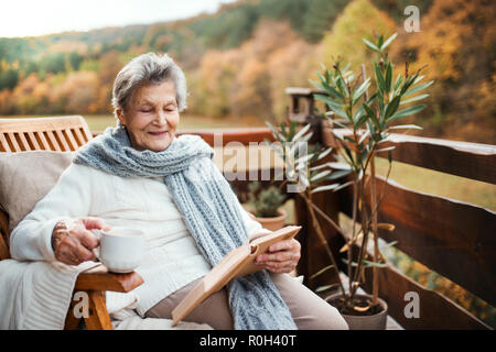 Eine ältere Frau liest Buch draußen auf einer Terrasse an einem sonnigen Tag im Herbst. Stockfoto