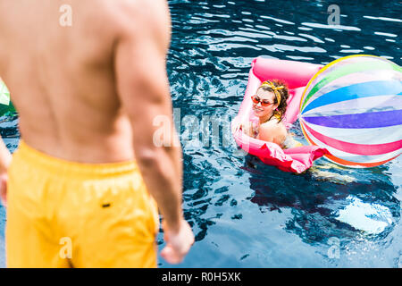 Zugeschnittenes Bild der Mann am Pool, während seine Freundin Schwimmen auf aufblasbare Matratze mit einem Ball im Pool Stockfoto