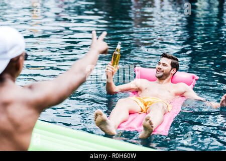 Lächelnder Mann mit Bier auf aufblasbare Matratze im Schwimmbad, während sein Freund gestikulierend durch die Finger am Pool Stockfoto