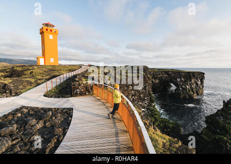 SNAEFELLSNES, ISLAND - August 2018: Frau im gelben Regenmantel Svortuloft beobachten Sonnenuntergang am Leuchtturm. Stockfoto