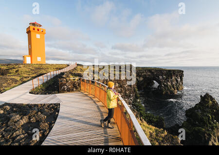 SNAEFELLSNES, ISLAND - August 2018: Frau im gelben Regenmantel Svortuloft beobachten Sonnenuntergang am Leuchtturm. Stockfoto