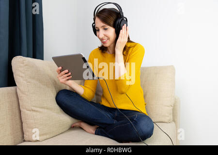 Schöne Frauen sitzen auf einem Sofa und hören mit Kopfhörern. E-Learning - Konzept. Stockfoto