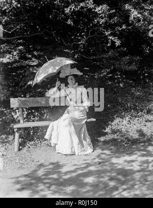 Ein Edwardianisches schwarz-weiß Foto zeigt eine junge Frau sitzt auf einer Bank unter einem Baum, in der feinen formelle Kleidung, während sie gleichzeitig ein Sonnenschirm über ihrem Kopf. Stockfoto
