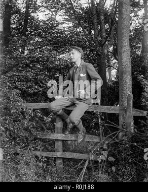 Edwardian schwarz-weiß Foto eines Jungen in Gap, Jacke gekleidet und verletzt und sitzt auf einem Zaun mit Wäldern im Hintergrund.