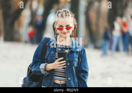 Hipster attraktive Mädchen mit dreads in Kopfhörer und die Brille Jeansjacke Kaffee trinken und gehen auf die Straße Stockfoto