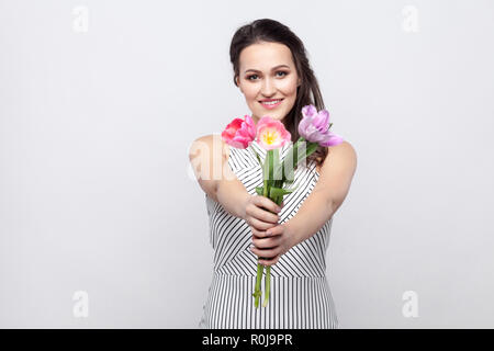 Porträt der jungen brunette schöne Frau mit Make-up in weiss gestreiftes Kleid, stehen und die Tulpen, die Kamera und die Pflege von toothy Lächeln. Stockfoto