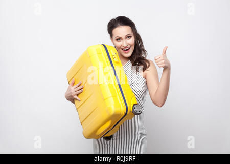 Glückliche junge Reisende in gestreiften Kleid Stehen und Halten der gelbe Koffer und zeigte mit Daumen hoch mit offenen Mund und bereit für den Urlaub. Innen- Studio Stockfoto