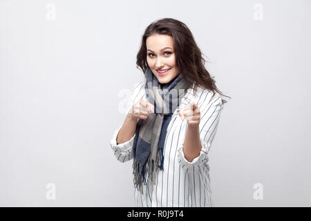 Portrait von lustigen glücklich schöne Brünette Frau in weiß gestreifte Jacke und blauer Schal stehen, zu Kamera zeigt mit toothy Lächeln. indoor Studio Stockfoto