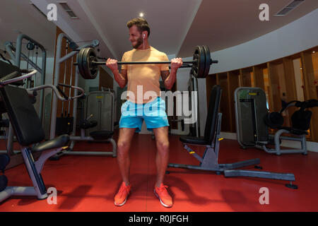 Jungen männlichen Erwachsenen tun Barbell bicep Curls in der Turnhalle beim Hören von Musik über Kopfhörer. Stockfoto