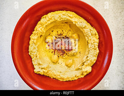Eine rote Schale hausgemachter Hummus gewürzt mit Paprika und Olivenöl über einem weißen Granit Tisch. Ansicht von oben. Stockfoto