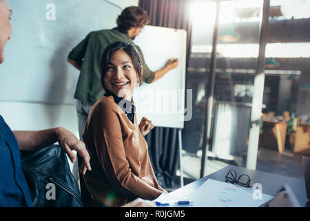 Lächelnde asiatische Frau, die mit einem Kollegen spricht, der in einer Geschäftspräsentation vorbeisitzt. Geschäftspräsentation im modernen Sitzungssaal. Stockfoto
