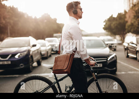 Geschäftsmann zu Fuß auf der Straße sein Fahrrad die Straße überqueren. Mann, office Bag und kabellose Kopfhörer gehen zu Office sein Fahrrad halten. Stockfoto