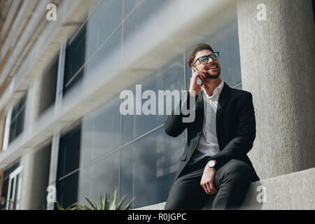Lächelnd mann Gespräch am Handy außerhalb eines Gebäudes sitzen. Mann eine Pause von der Arbeit sprechen auf Handy vor seinem Büro sitzen. Stockfoto